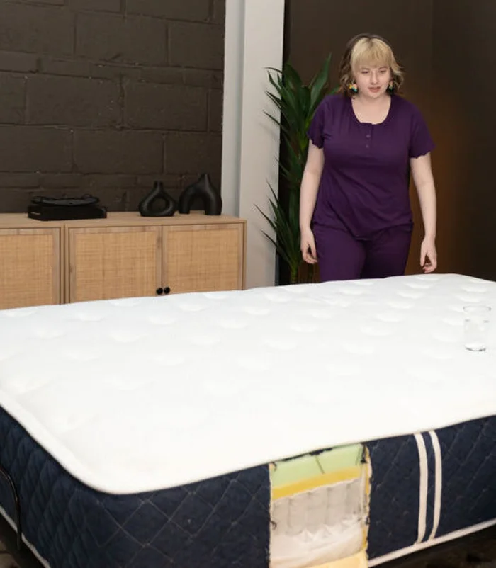 A woman stands next to the Brooklyn Bedding Signature Hybrid mattress, which has a cup of water balanced on its surface