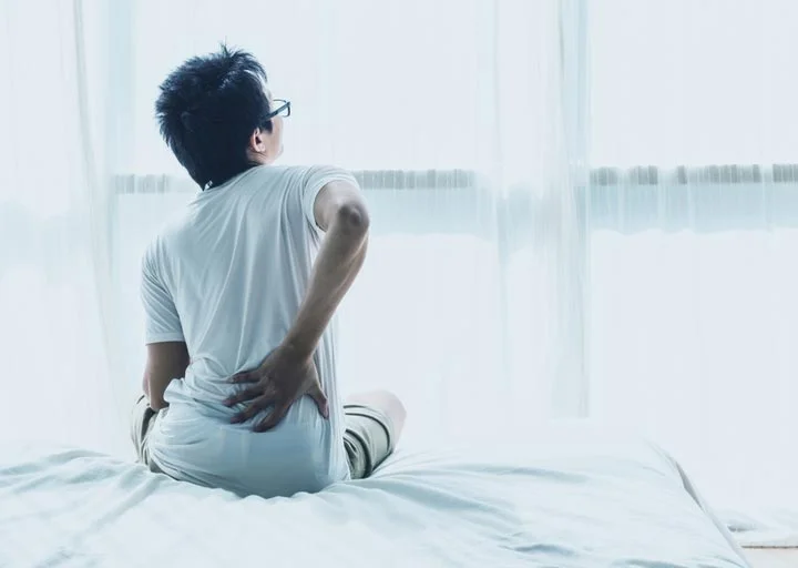 How to Sleep with Back Pain A man sitting on a bed clutches his lower back, showing that it's in pain