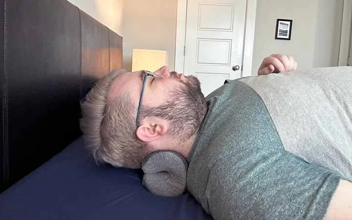 A wide shot of a man resting his head and neck on the NekGenic Neck Pillow.