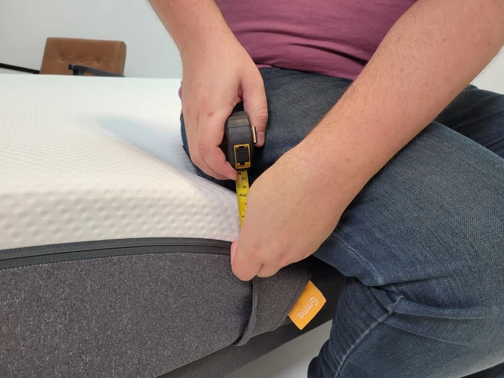A man sits on the edge of the Emma mattress with a tape measure displaying 3 inches of sagging
