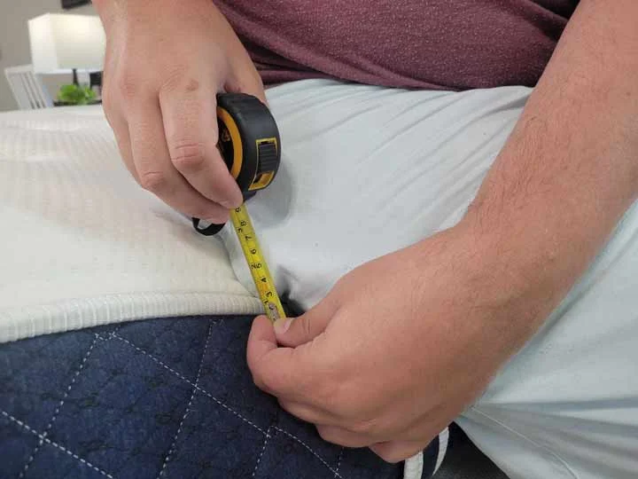 A close up image of a man holding a tape measure, showing how deep the sinkage of the edge is when sitting along the edge of the Brooklyn Bedding Signature.