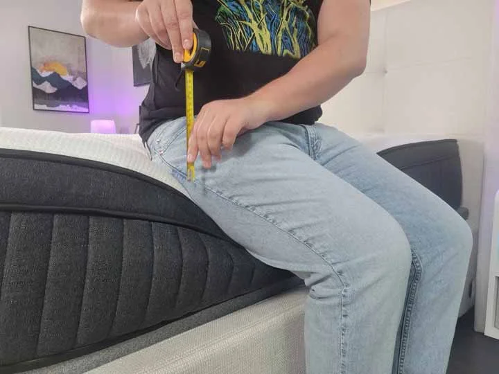 A man sits on the edge of the Titan Plus Luxe. He has a tape measure to show how much the edge of the mattress is dipping.