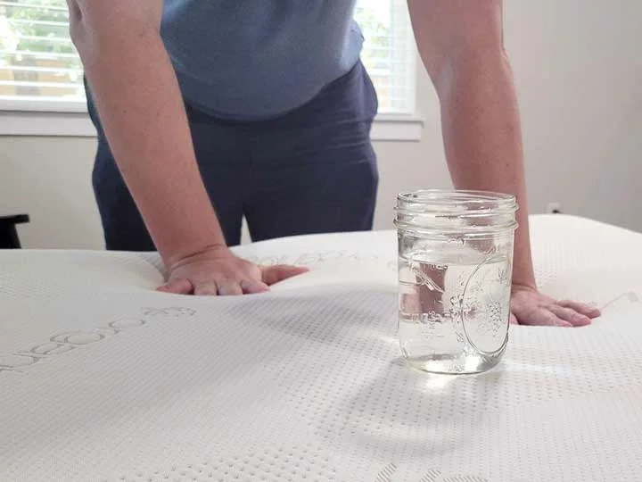 An image of a glass of water on top of the Saatva Contour5. Hands press into the area round the glass.