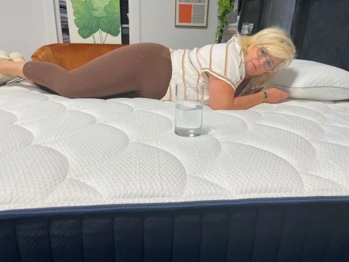 A woman tests motion isolation on the DreamCloud Memory Foam mattress with a glass of water