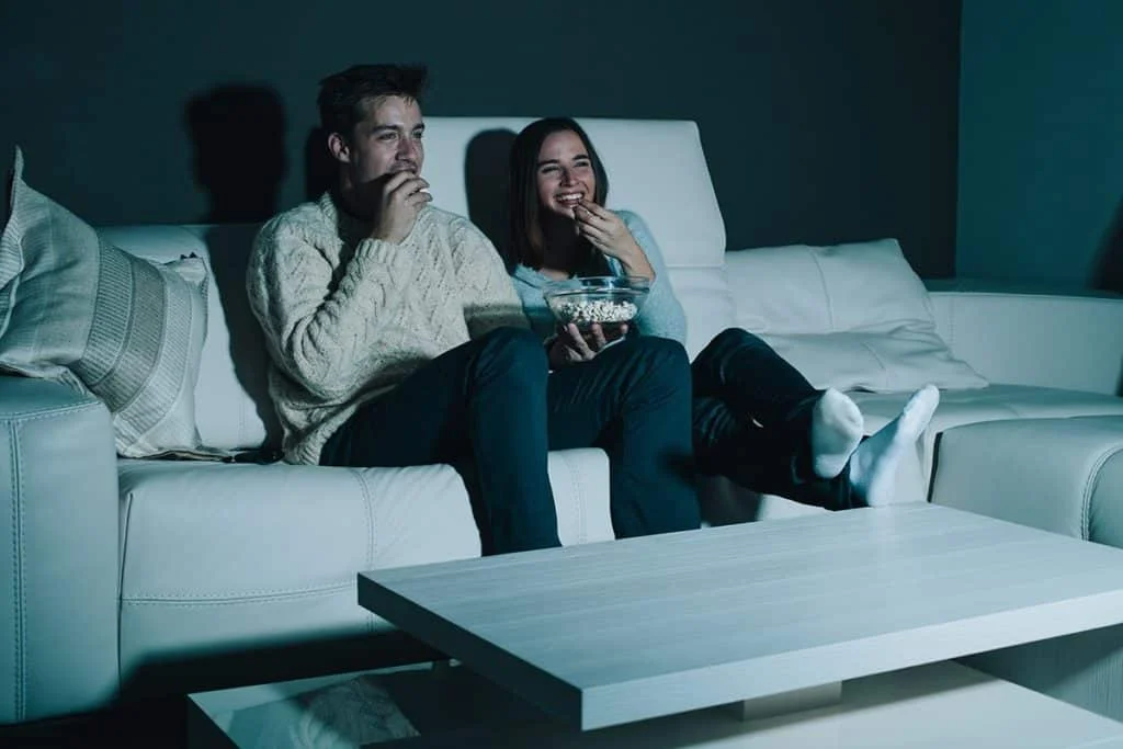 How Binge Watching TV Affects Sleep A man and woman watch television on a couch while eating popcorn
