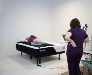 A man lies on a pressure-sensitive mat on the Nectar Classic mattress while a woman in purple pajamas looks on