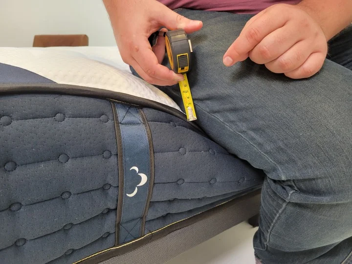 A man uses a tape measure to measure sinkage at the edge of the DreamCloud mattress while sitting on it
