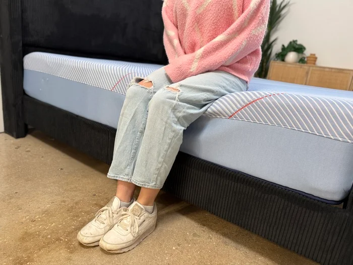 woman in a pink sweater sitting on the edge of a sealy posturepedic hybrid mattress